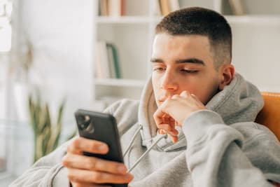 A teenage boy in a purple hoodie and headphones sits on a sofa, using his smartphone in a bright, cozy living room. The scene evokes a sense of modern adolescence and connectivity.

